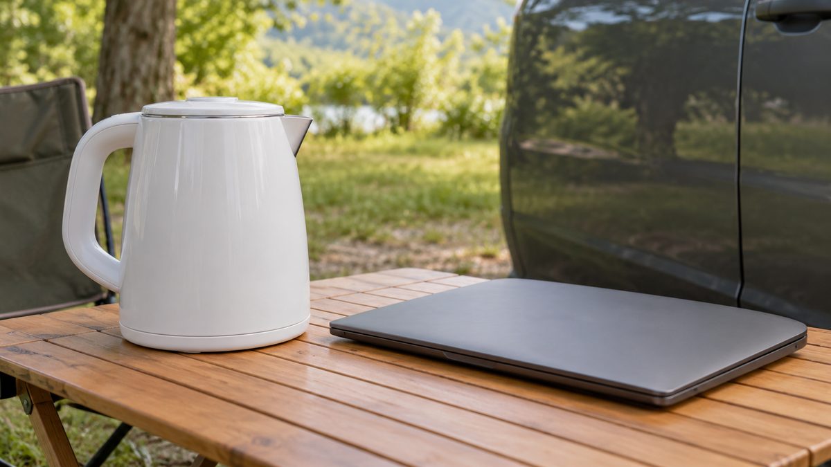 An unplugged travel kettle and closed laptop rest on a camping table beside a parked car door.