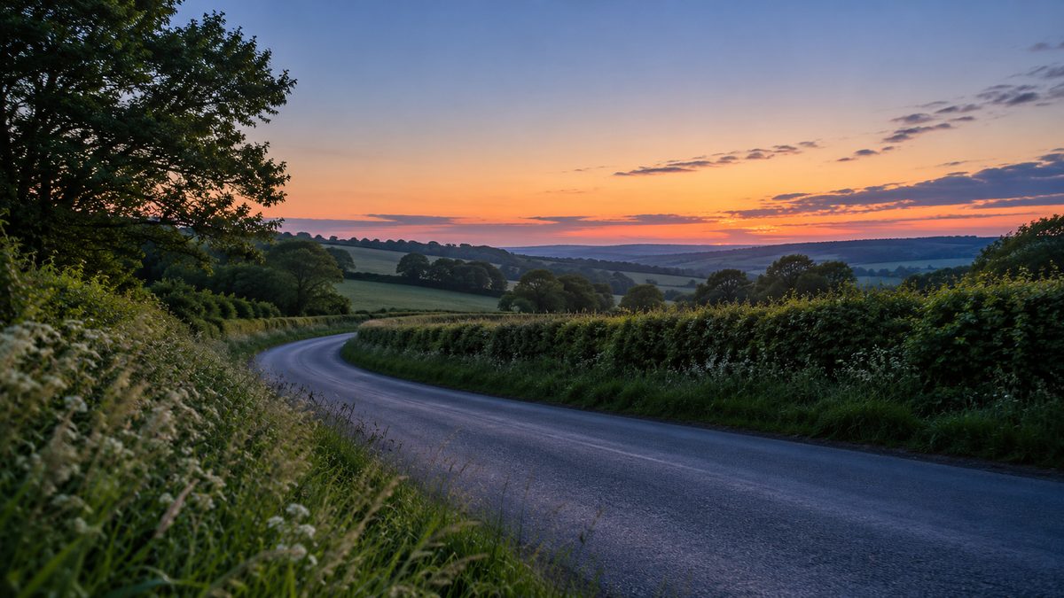 An empty country road curves between green hedgerows beneath a soft orange and blue evening sky.