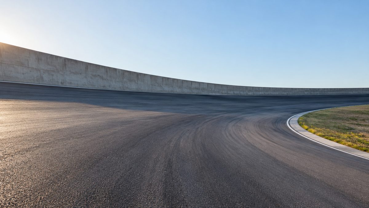 An empty banked test-track curve rises beside a plain concrete wall beneath a clear morning sky.