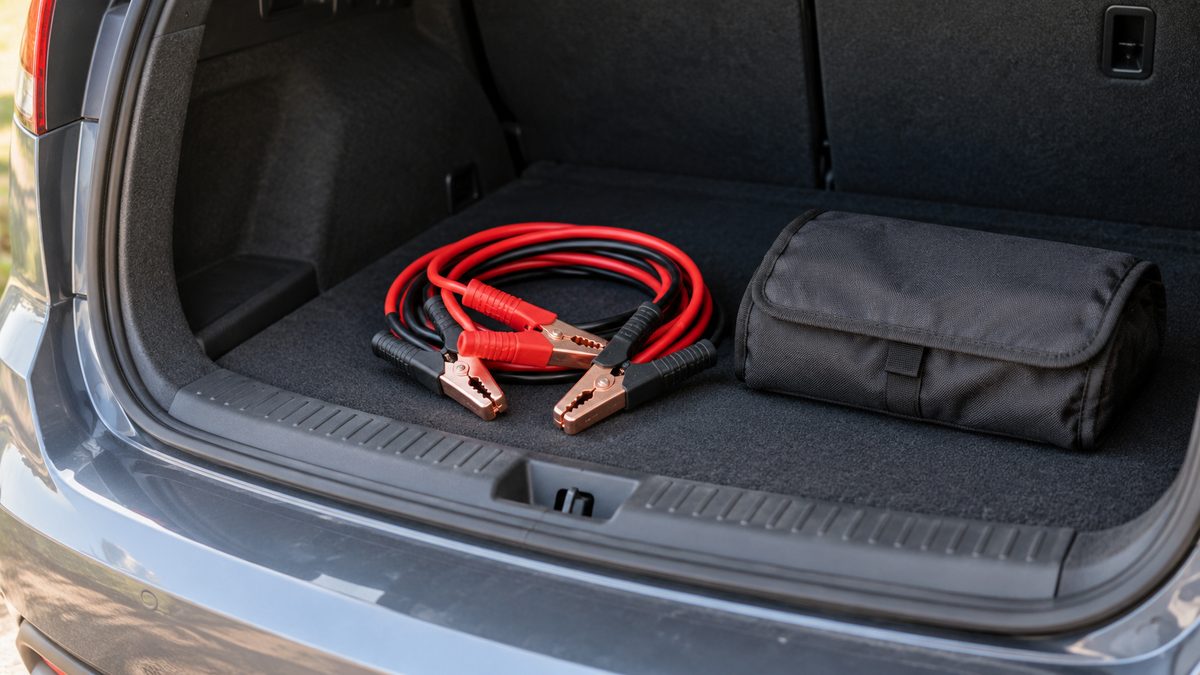 Red and black jumper cables lie coiled beside a plain fabric pouch on the mat inside an open car boot.