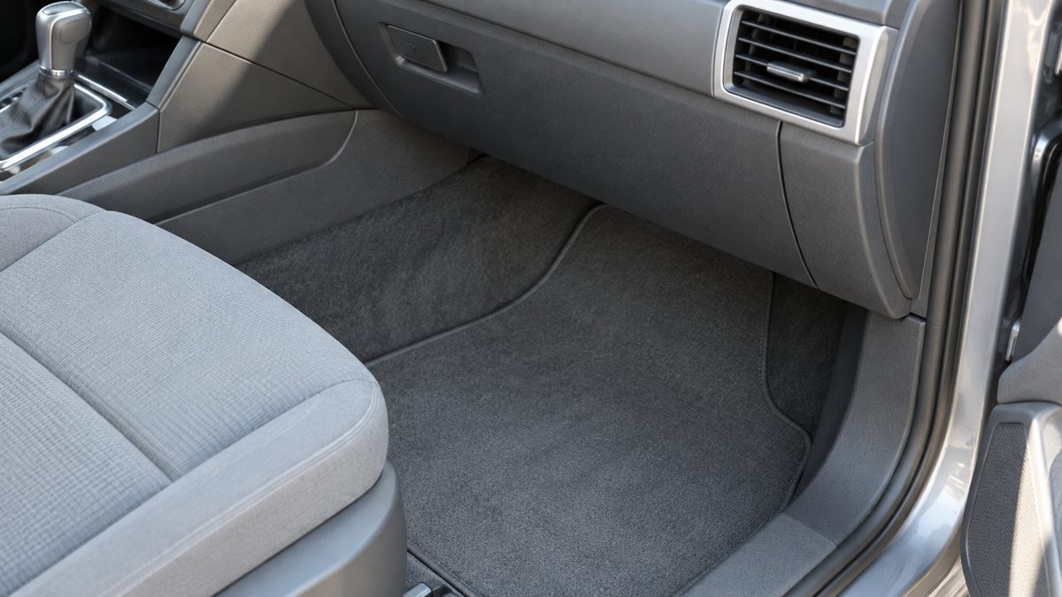 A clean passenger footwell and plain ventilation outlet sit beneath a car dashboard in soft daylight.