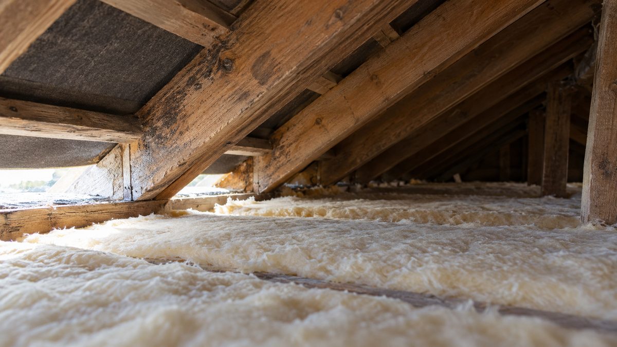 Weathered rafters frame a low loft space with pale insulation and daylight entering near the eaves.