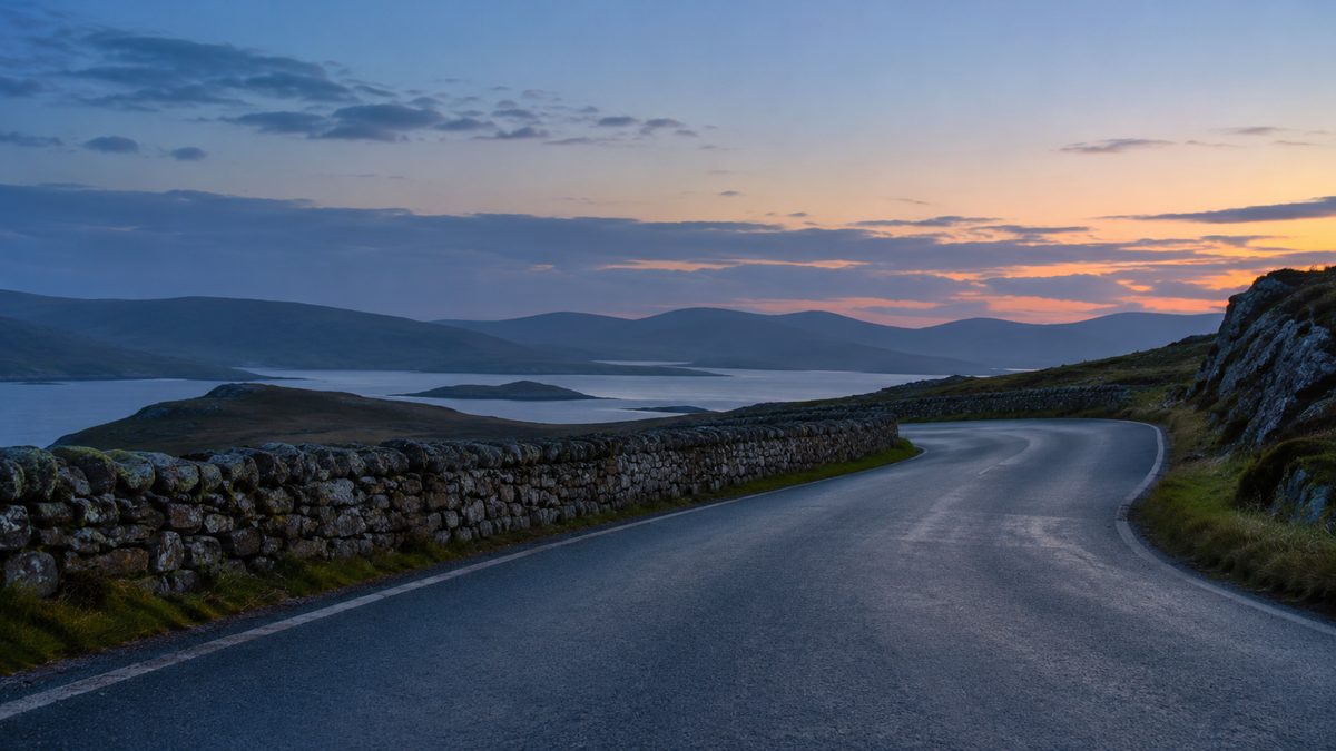 A quiet empty road curves beside a low stone wall toward distant hills in soft blue evening light.