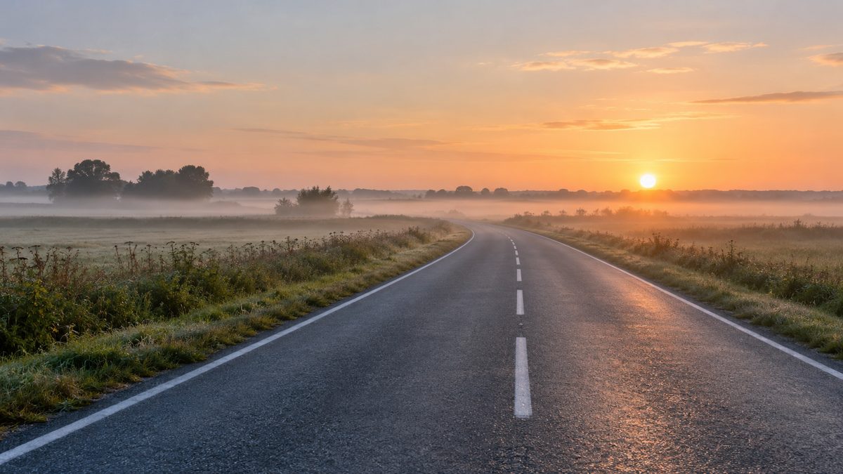 A quiet empty road crosses a flat rural landscape beneath low mist and soft dawn light.